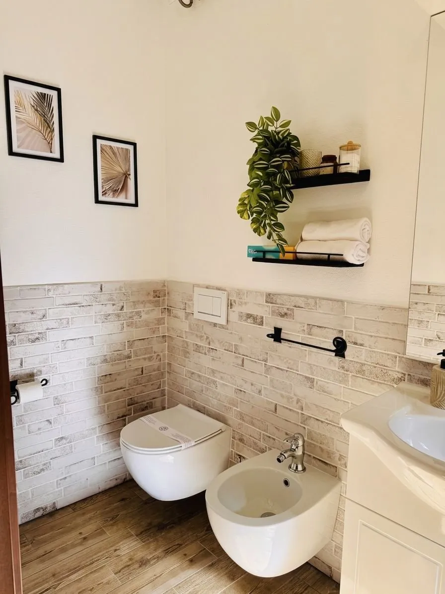Bagno completo della suite a Cagliari: WC e bidet sospesi, lavabo con mobile shaker bianco, rivestimento effetto mattoncini, stampe foglie di palma, mensole nere con piante e amenities.