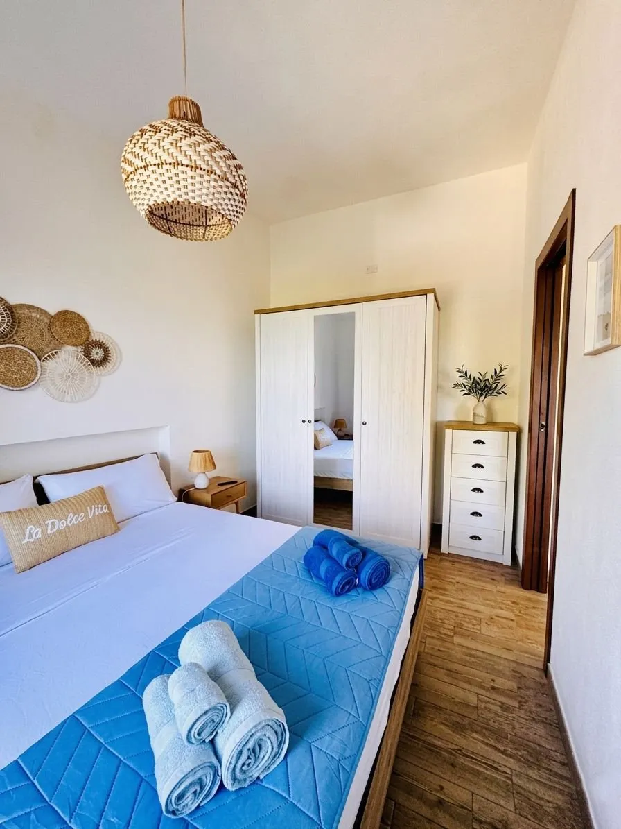 Camera matrimoniale stile boutique mediterraneo: armadio bianco con specchio, cassettiera con vaso d'ulivo, lampadario in vimini intrecciato, dettagli artigianali sardi.