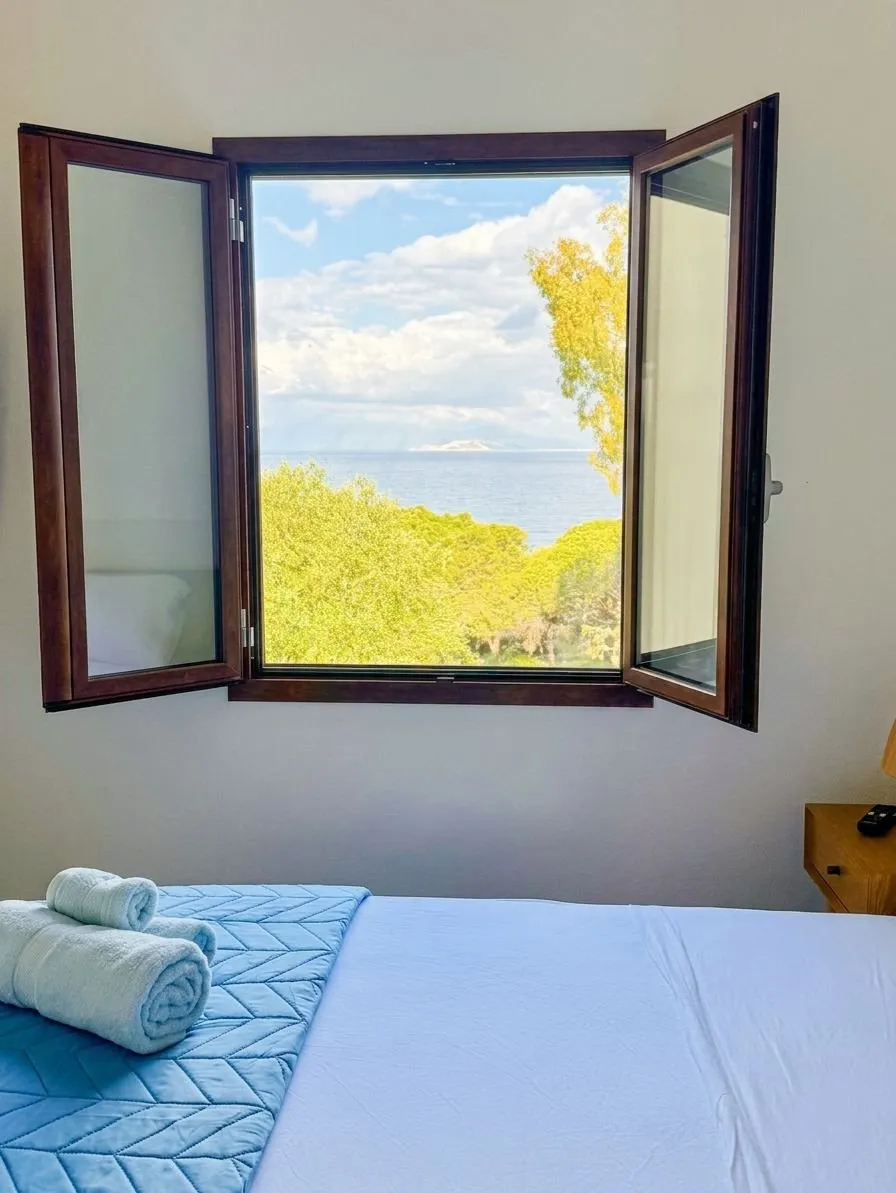 Camera da letto della suite a Cagliari con finestra in legno mogano spalancata sul Golfo: mare blu intenso, profilo di un'isola all'orizzonte, pineta mediterranea, il blu è la prima cosa che vedi al risveglio.