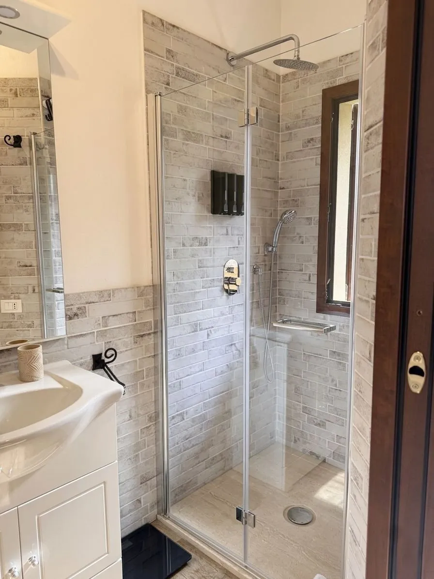 Doccia walk-in del bagno: cabina in vetro, soffione effetto pioggia cromato, piatto doccia in travertino, finestra in legno mogano per luce naturale.