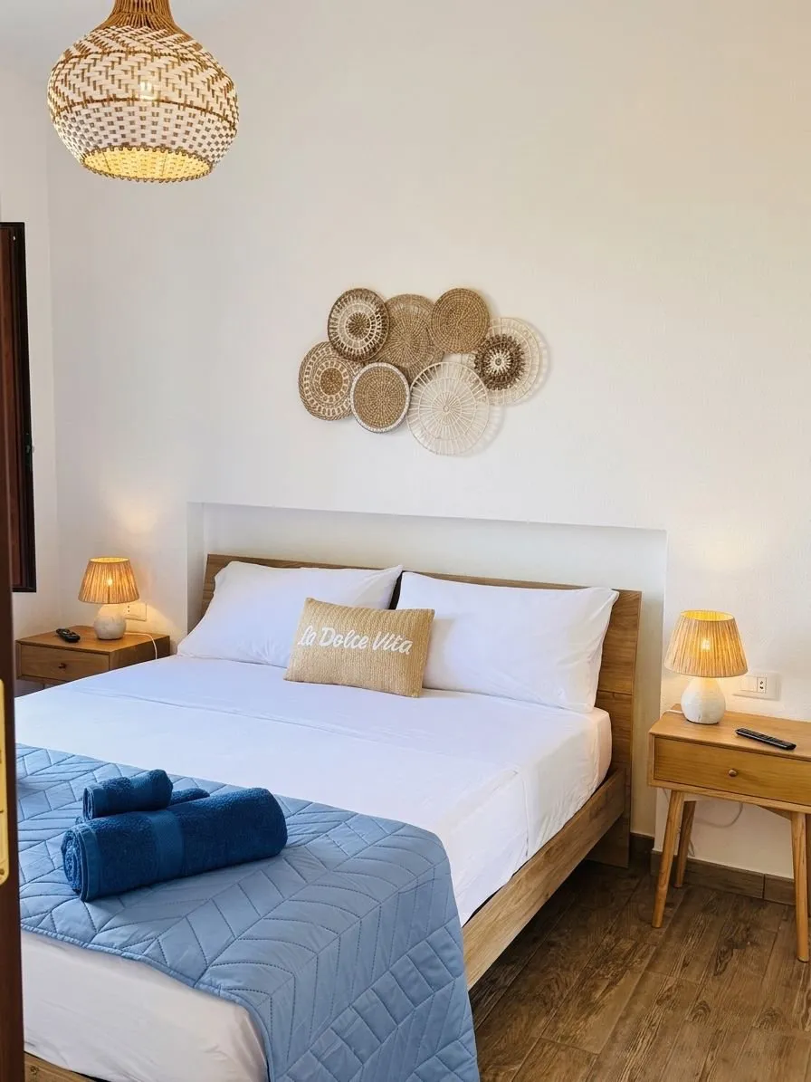 Camera matrimoniale della suite a Cagliari, vista frontale: letto king-size con cuscino 'La Dolce Vita', lampade da tavolo in rattan accese, decorazione a parete con cestini in vimini.