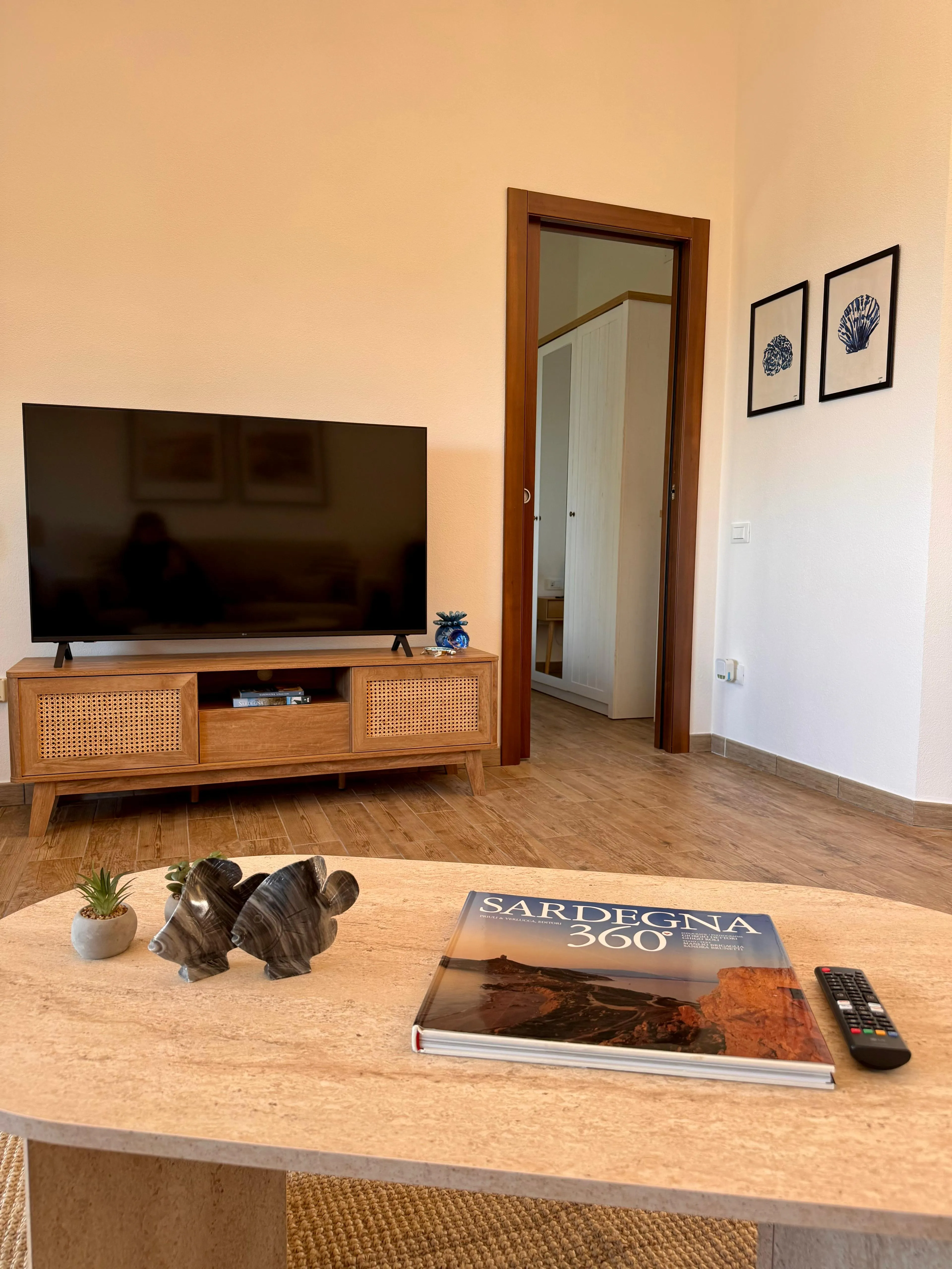 Living della suite a Cagliari, prospettiva grandangolare: tavolino in travertino con libro fotografico 'Sardegna 360°', scultura decorativa, mobile TV in legno chiaro con ante in paglia di Vienna, stampe coastal con conchiglie blu navy.