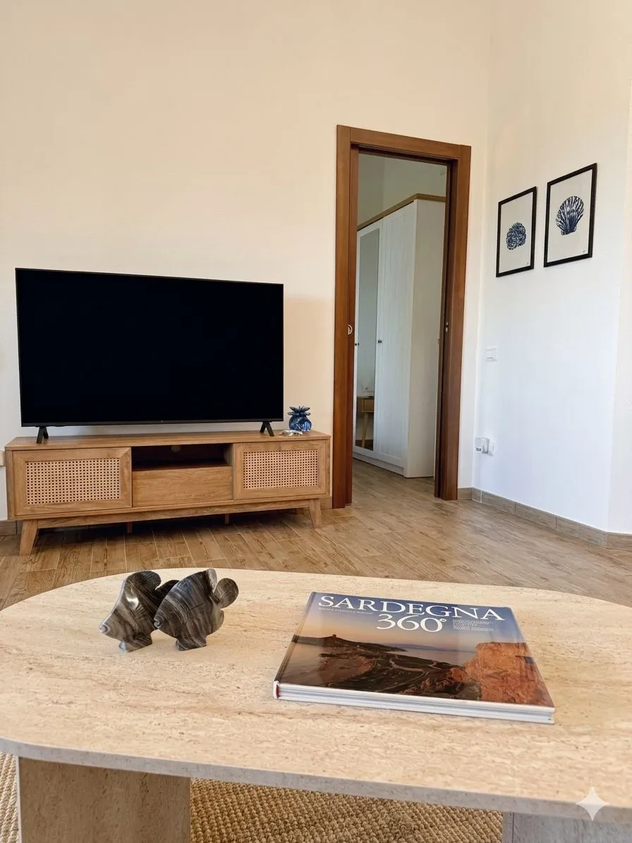 Zona TV del soggiorno: Smart TV su mobile in legno chiaro con ante in paglia di Vienna, libro fotografico Sardegna 360° e scultura decorativa sul tavolino in travertino.