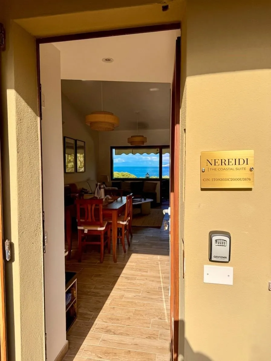 Ingresso della suite Nereidi a Cagliari: targa dorata 'NEREIDI - The Coastal Suite' con codice CIN visibile, porta aperta che inquadra il soggiorno e la vista mare in fondo.