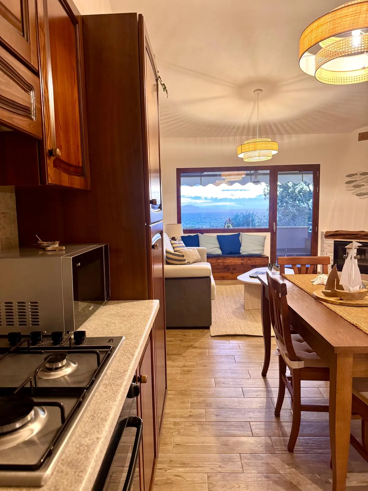 Open space della suite a Cagliari: la cucina in legno massello apre sul tavolo da pranzo e sul soggiorno con divano e cuscini blu navy, fino alla porta-finestra che inquadra il mare blu del Golfo. Tutto in un colpo d'occhio.