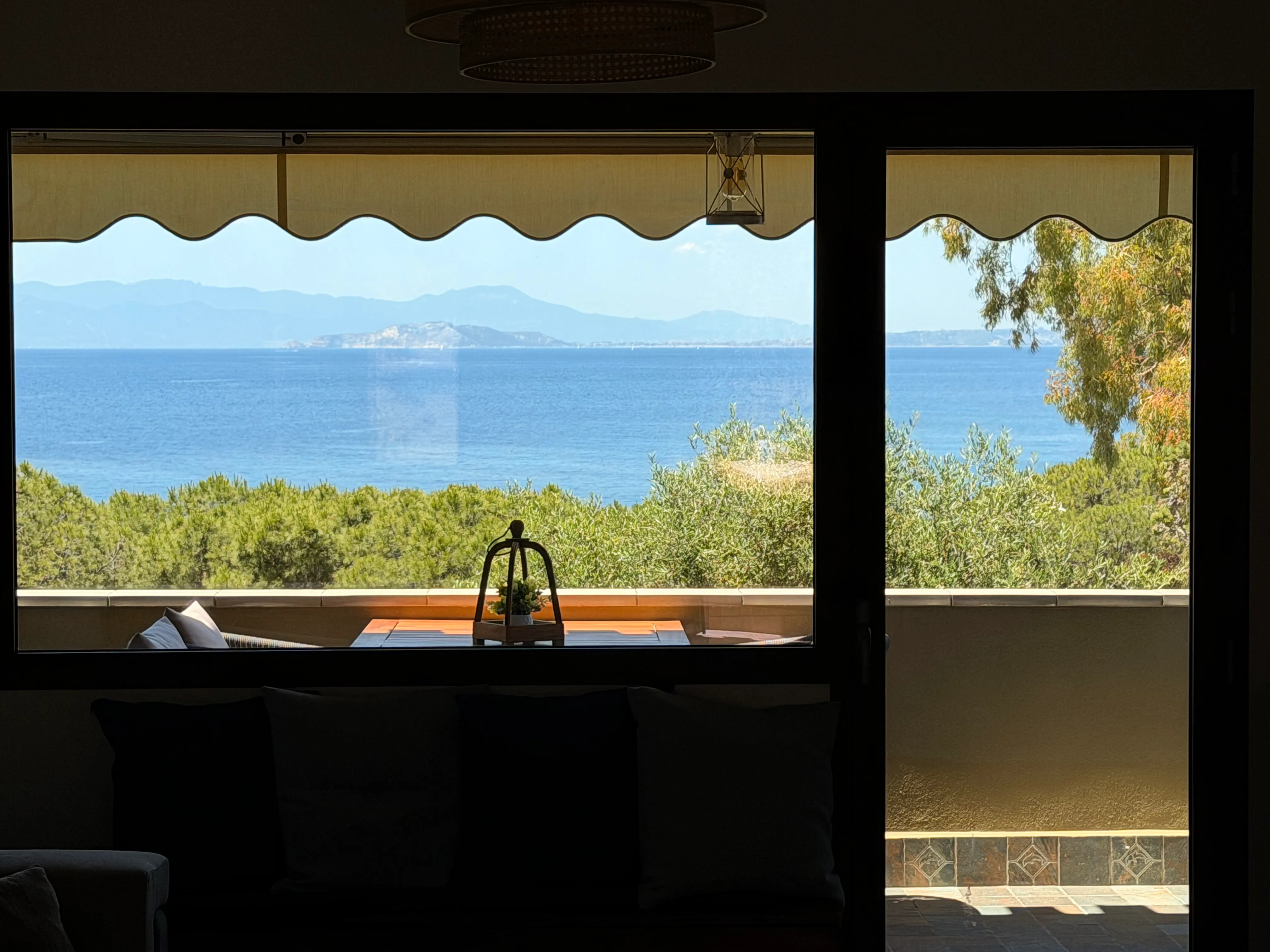 Vista panoramica dal salone di The Coastal Suite a Cagliari: finestra e porta-finestra con tenda dal bordo ondulato e lanterna decorativa, divano con cuscini, profilo costiero del Golfo di Cagliari oltre la vegetazione mediterranea.