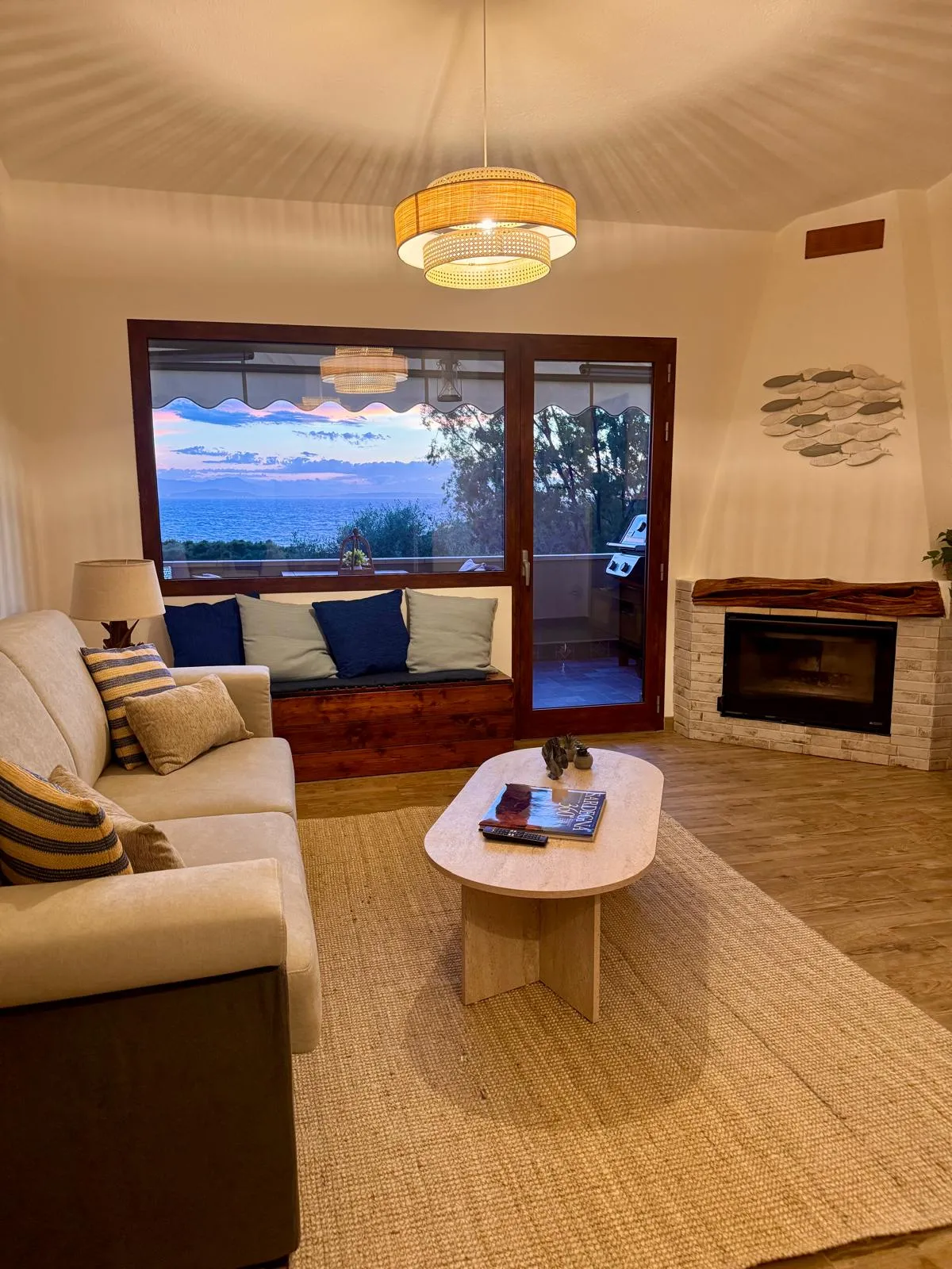 Soggiorno della suite al crepuscolo: lampada in rattan accesa con luce calda, divano bianco con cuscini blu navy, porta-finestra che inquadra il cielo rosa lilla del tramonto sul Golfo di Cagliari, libro 'Sardegna 360°' sul tavolino in travertino.