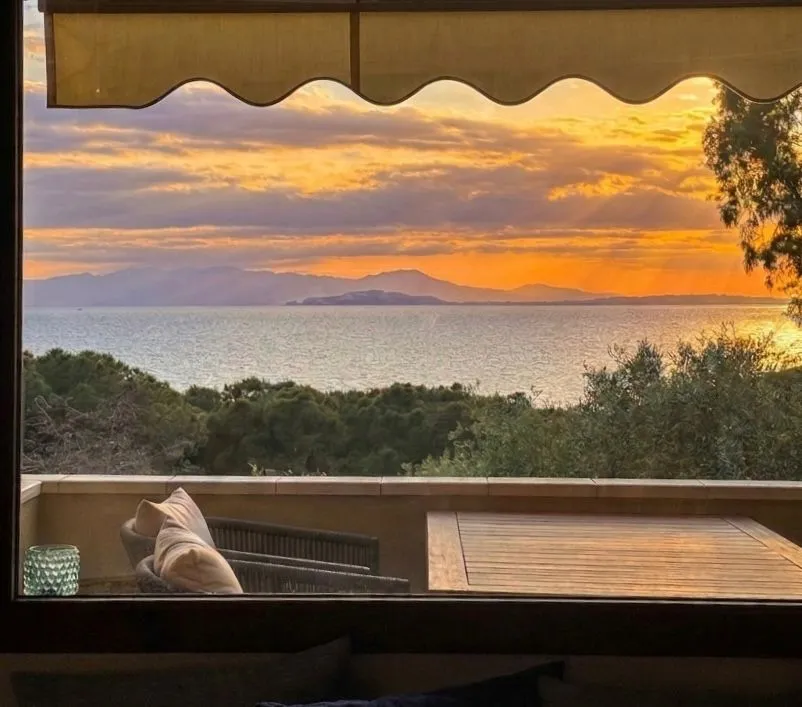 Tramonto dalla terrazza della suite a Cagliari: divano outdoor con cuscino chiaro in primo piano, tavolo in legno, cielo arancione con raggi tra le nuvole sul Golfo di Cagliari, profilo delle montagne all'orizzonte.