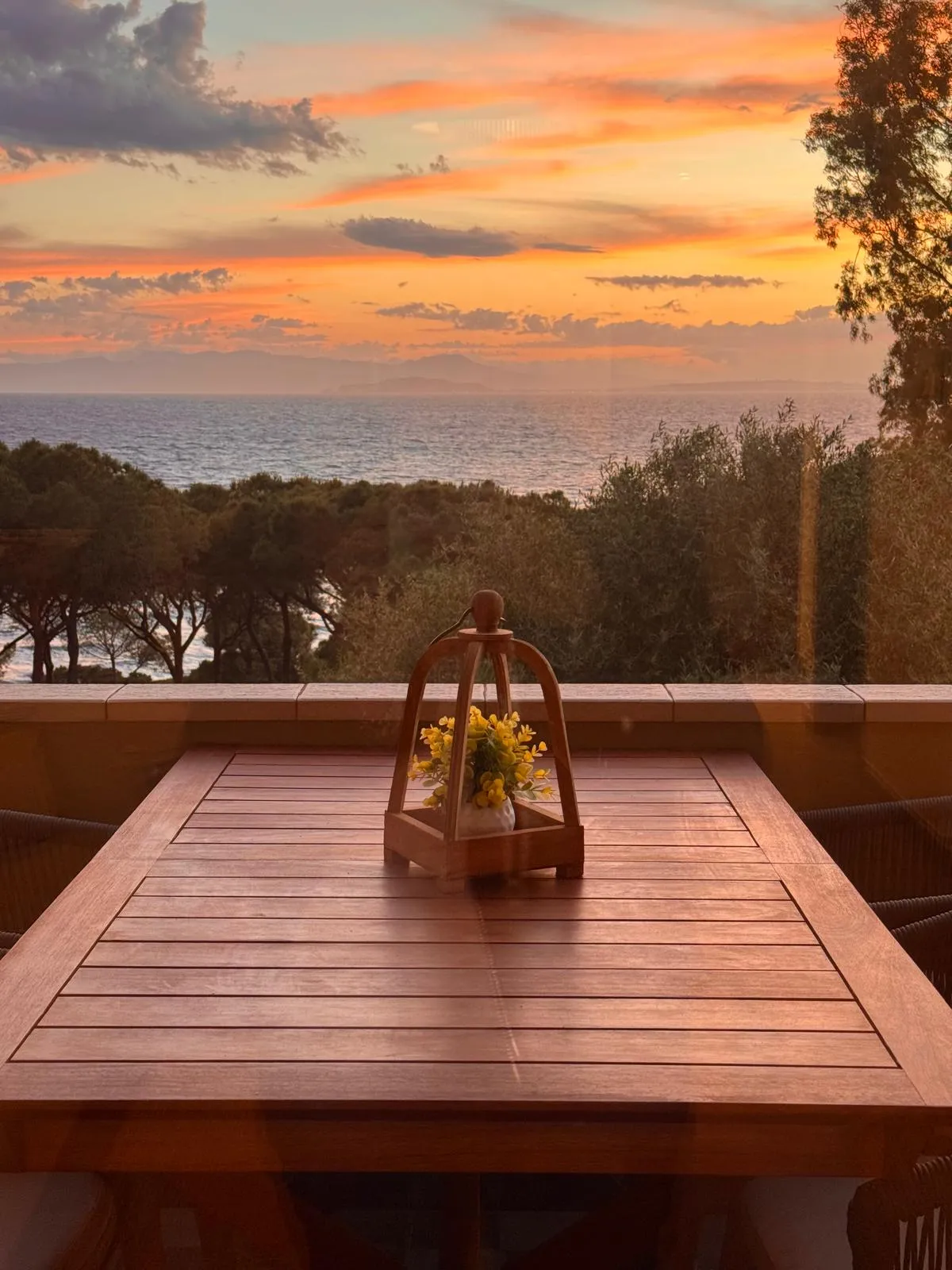 Terrazza della suite al tramonto sul Golfo di Cagliari: tavolo in legno con lanterna decorativa nera come punto focale, sedie in corda blu navy, cielo arancione e rosa pesca, tenda con bordo ondulato.