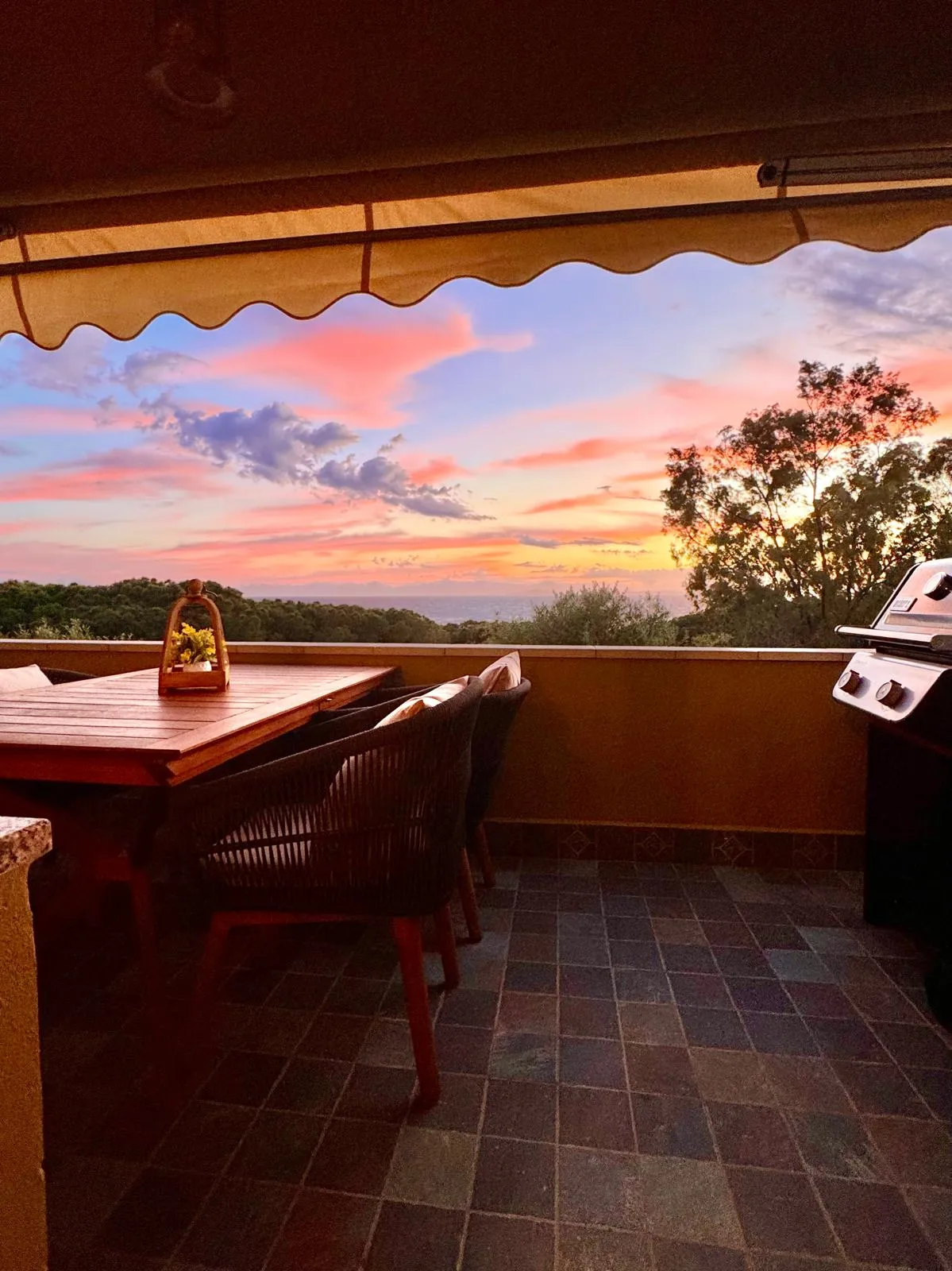 Terrazza della suite a Cagliari al tramonto: tavolo da pranzo outdoor in legno con sedie in corda blu navy, lanterna decorativa, barbecue Weber a destra, cielo rosa pesca sul Golfo di Cagliari, vegetazione mediterranea.