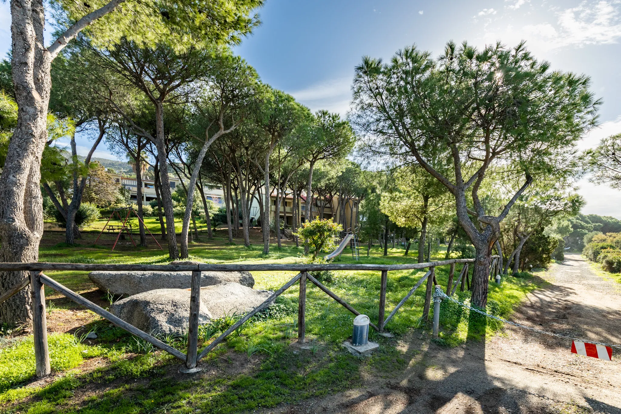 Area centrale del villaggio Marina delle Nereidi: steccato in legno rustico, grandi pini marittimi su prato verde, sentiero sterrato e case basse beige sullo sfondo, cielo limpido.