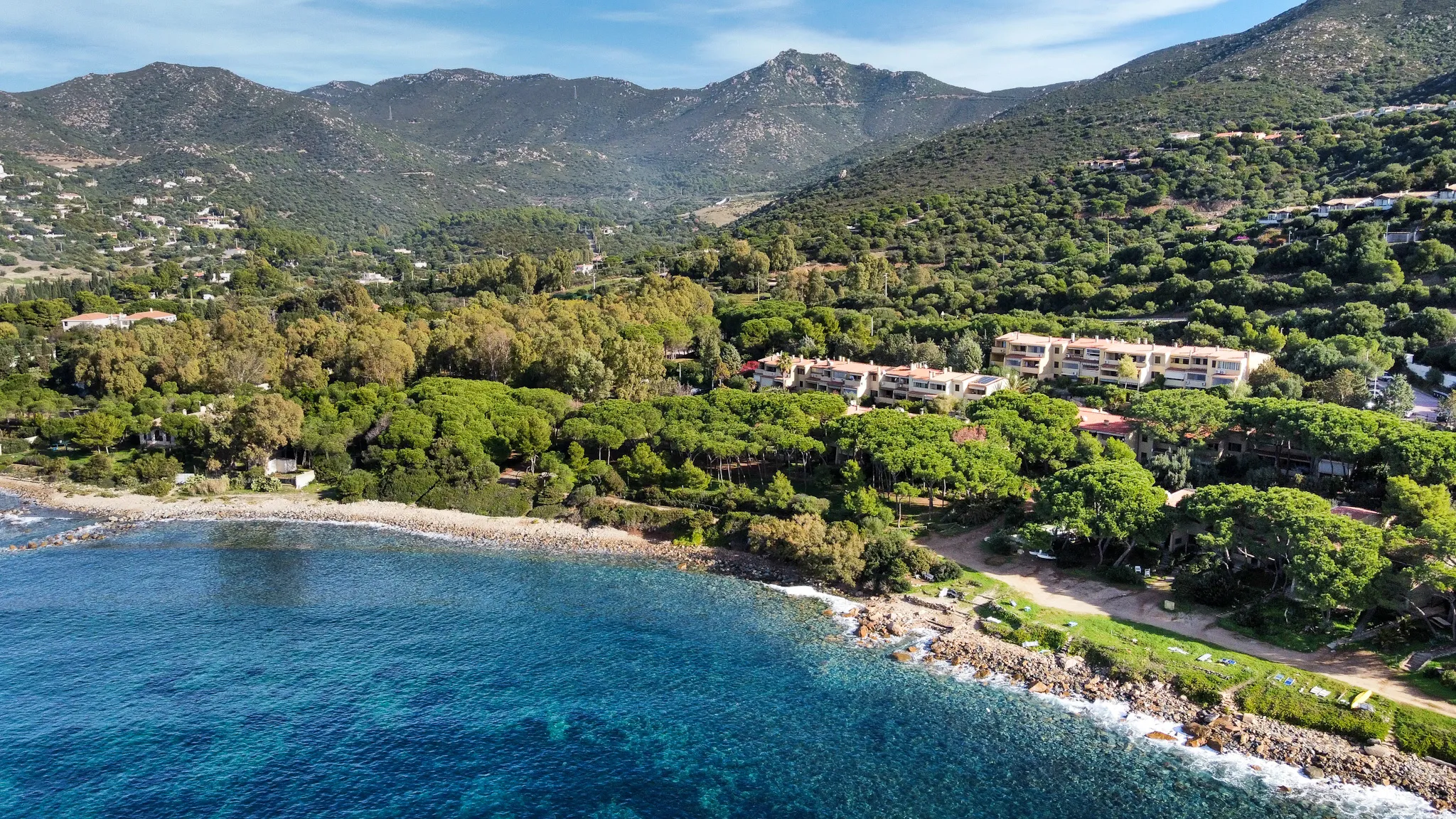 Vista aerea drone di Marina delle Nereidi: la costa rocciosa con il mare turchese del Golfo di Cagliari, la pineta che avvolge le case beige del villaggio, le colline e le montagne dell'entroterra sardo.