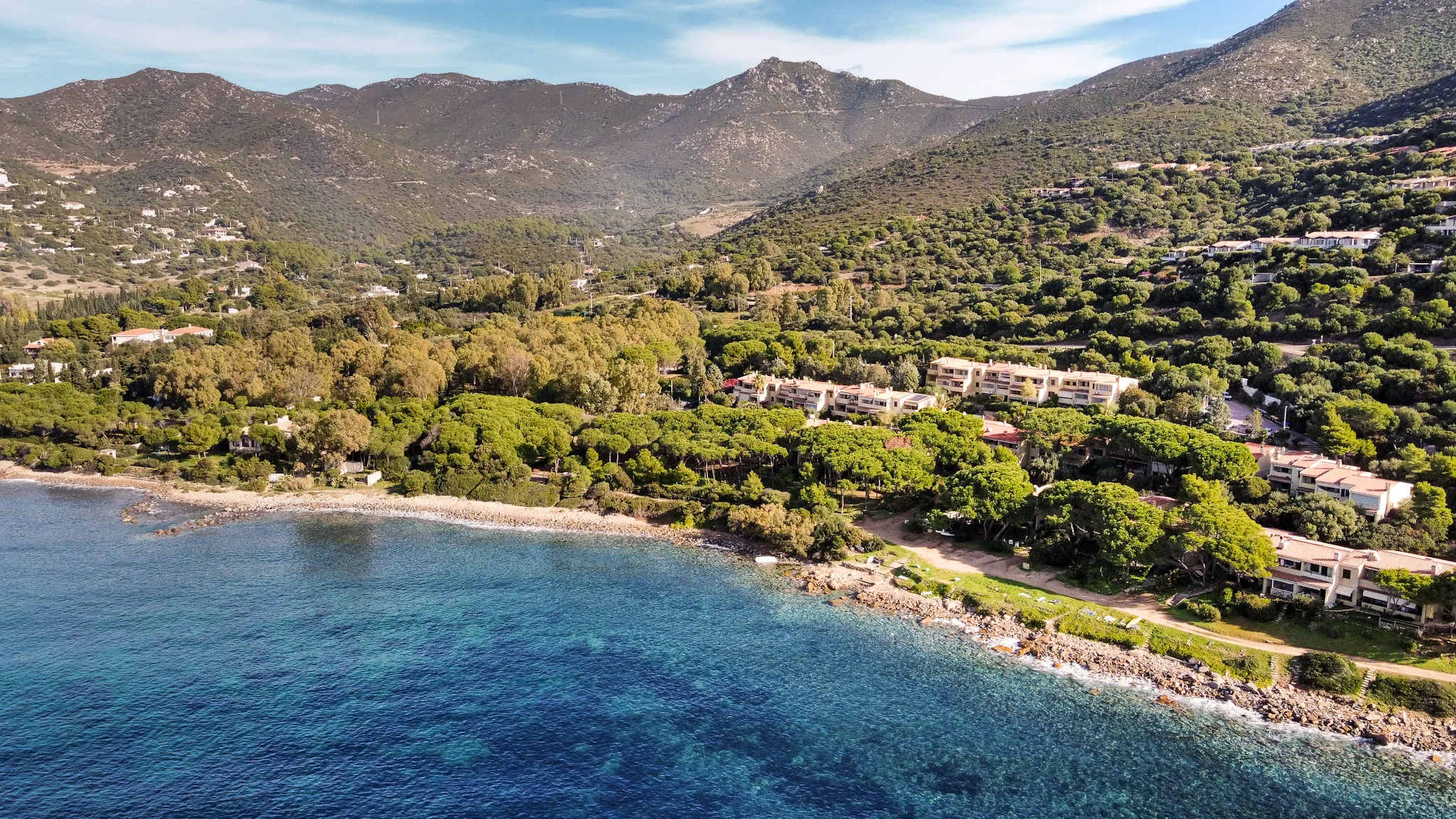 Vista aerea drone del villaggio Marina delle Nereidi: la costa rocciosa con il mare turchese in primo piano, la pineta mediterranea che avvolge le case beige del villaggio, le colline e le montagne dell'entroterra sullo sfondo.