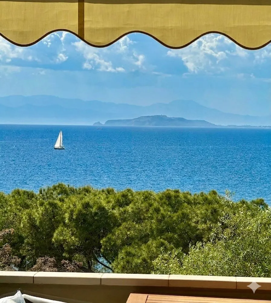 Vista mare dalla suite a Cagliari: una vela bianca taglia il blu del Golfo, l'isola si staglia all'orizzonte, la pineta in primo piano incornicia il quadro sotto la tenda con bordo ondulato.