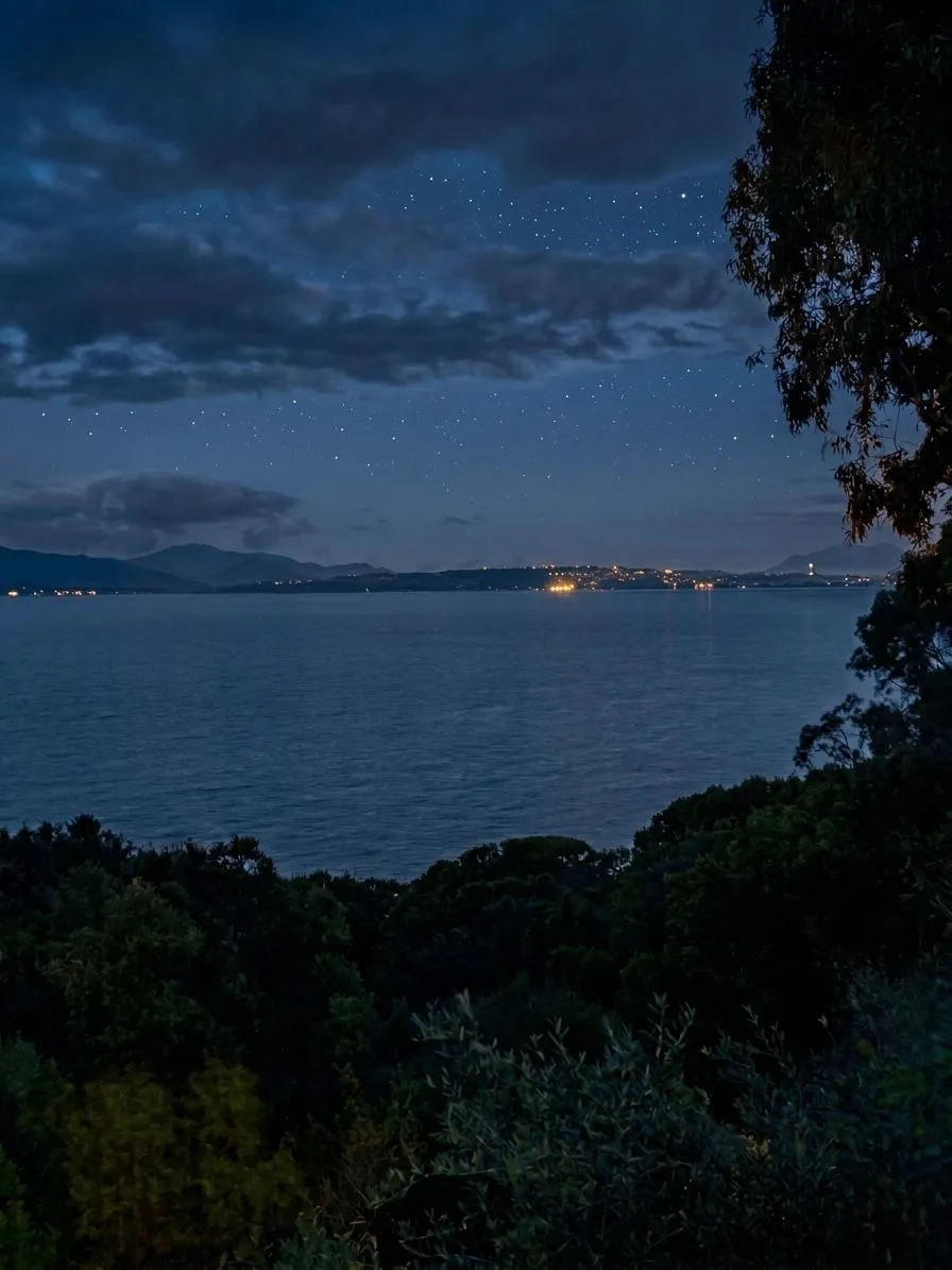 Vista notturna dal balcone della suite a Cagliari: cielo stellato sul Golfo, le luci di Cagliari all'orizzonte - da Marina Piccola al Castello - riflesse sul mare calmo, silhouette della vegetazione mediterranea in primo piano.